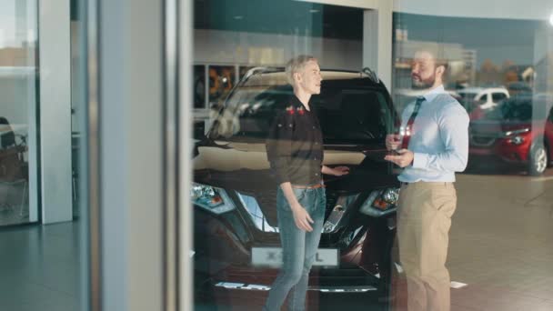 Dospělá zákaznice a usměvavý prodejce aut stojí v interiéru autosalonu. Usmívající se přátelský muž prodejce aut s tabletem v ruce mluvit se zákaznicí v černé košili, která chce koupit auto. — Stock video