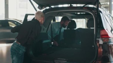 Araba satıcısı otomobil galerisinde müşteri arabasını gösteriyor. Ayrıntılar arabanın bagajında. Gülümseyen dost canlısı araba satıcısı siyah gömlekli bir kadın müşteriye arabanın içini gösteriyor.