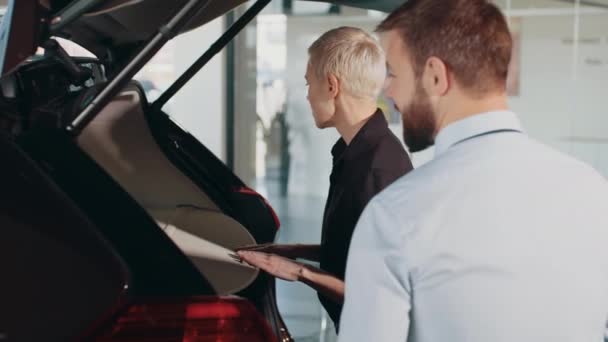 Femme choisir une voiture chez un concessionnaire automobile et d'inspecter le coffre. Coffre d'une voiture neuve dans un concessionnaire automobile. Femme d'affaires avec un vendeur examine le grand coffre d'une nouvelle voiture en concession — Video