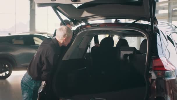 Begreppet människor och transport. Bilförsäljare visar kundbil i bilhandlarnas showroom. En uppmärksam kvinnlig köpare och manlig säljare studerar detaljerna i den öppna bakluckan på bilen. — Stockvideo