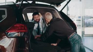 Gülümseyen dost canlısı araba satıcısı siyah gömlekli bir kadın müşteriye bagajın içini gösteriyor. Müşteri ve gülümseyen erkek araba satıcısı, otomobil kuaförünün önünde bagajı açık duruyor..