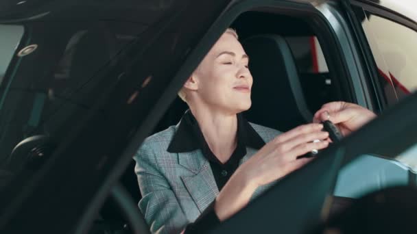 Gorgeous blond woman who is sat in car in car dealership smiling while receiving car keys to her new automobile in an automotive dealership salon. Woman buying auto. Concept of people and transport. — стоковое видео