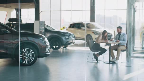 European female client signing papers with salesman in dealership store. Visiting car dealership. Handsome sales manager is smiling while beautiful client is signing papers and gets the keys — Stock Video