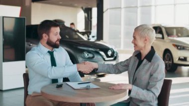 Çekici bir bayan müşteri araba satın alma belgesi okuyor ve otomobil kuaföründe torbacıyla masada otururken el sıkışıyor. Galeriden yeni araba alan bir kadın. Yeni ulaşım kavramı.