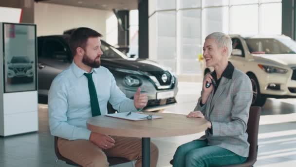 Mujer hermosa que se sienta en coche en concesionario de coches sonriendo mientras recibe las llaves de su coche nuevo en concesionario. Vendedor entregando la llave del coche a un cliente. Mujer comprando auto nuevo. Compra de auto nuevo — Vídeos de Stock