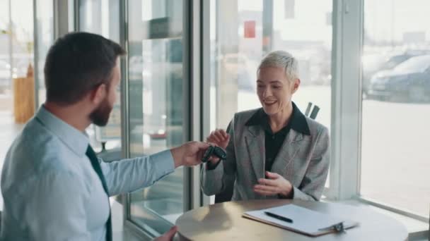 Smiling satisfied middle aged woman sitting at the table with car seller, shaking hands with him and taking car keys. Woman buying auto. Concept of people and transport. — стоковое видео