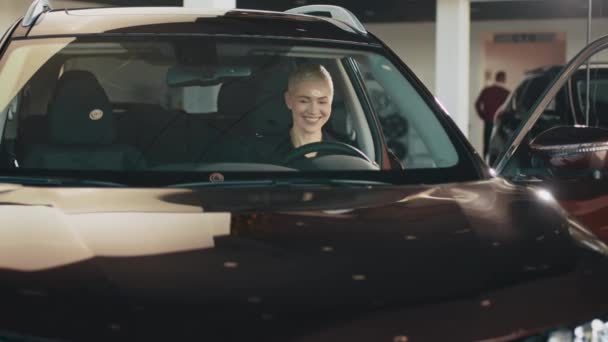 Uma mulher a conduzir um carro novo. Retrato de motorista mulher no carro. Mulher sorrindo em seu carro novo em um showroom. Mulher dirigindo um novo carro elétrico de luxo em uma concessionária de carros. A menina mostra emoções em seu novo carro. — Vídeo de Stock