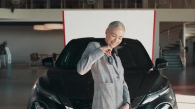 Güzel bir kadın araba galerisinde yeni arabanın yanında dururken elinde bir anahtar tutuyor. Kamera karşısında gülümseyen ve anahtarlarını gösteren olumlu bir kadın. Galeride yeni aracın yanında duruyor..