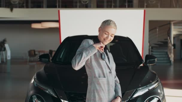 Beautiful woman is holding a key while standing near the new car in car dealership. Positive female in smiling for camera and demonstrating keys while standing near new vehicle in showroom. — Vídeo de stock