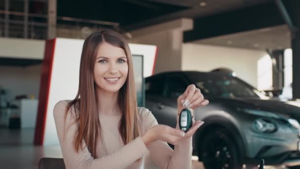 Mujer positiva en ropa casual inteligente sonriendo para la cámara y demostrando las teclas mientras está de pie cerca del nuevo vehículo en la sala de exposición. Joven alegre mostrando su nueva llave del coche en la concesionaria. — Vídeos de Stock