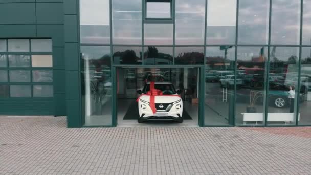 New white luxury car with big red bow riding out of modern auto salon. Expensive purchase of buyer. Concept of transport, surprise and selling. Aerial view of a new car with a bow — Stock Video