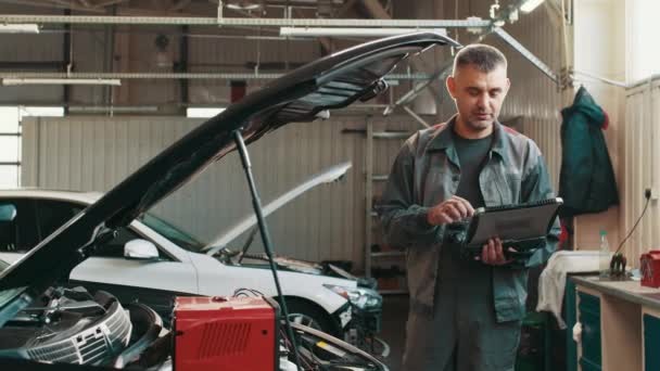 Uśmiechnięty kompetentny mężczyzna w średnim wieku robiący diagnostykę samochodu z laptopem w warsztacie. Auto serwis, naprawa, konserwacja koncepcja. Zamknij za pomocą tabletu cyfrowego do diagnostyki samochodu. — Wideo stockowe