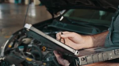 Otomobil servisi, bakım konsepti. Ellerinizi kapatın. Araba elektrikçisi araba tamir ediyor. Tamirci motor denetimlerini kaydetmek için dizüstü bilgisayar kullanır iş sırasında ayrıntılı bilgi toplar.