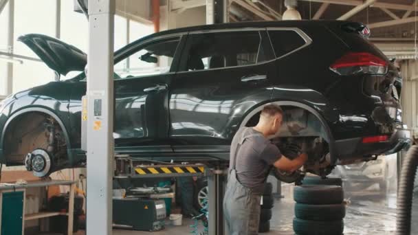 Car service, repair and maintenance. Professional car mechanic man working under lifted car in auto repair service. Bearded man doing auto repair with his hand near car wheel of raised up car — стоковое видео