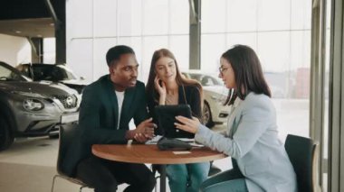 Araba kuaföründe çok ırklı bir çiftle çalışırken dijital tablet kullanan beyaz kadın. Mutlu adam ve kadın otomobil alırken yetkili yöneticiyle konuşuyor. Otomobil galerisindeki satıcıyla konuşuyorum.