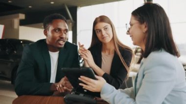 Modern kuaförde kadın satıcılarla otururken araba satın alma sözleşmesi imzalayan çok ırklı mutlu çift. Müdür galeride alıcılarla çalışırken dijital tablet kullanıyor. Otomatik iş