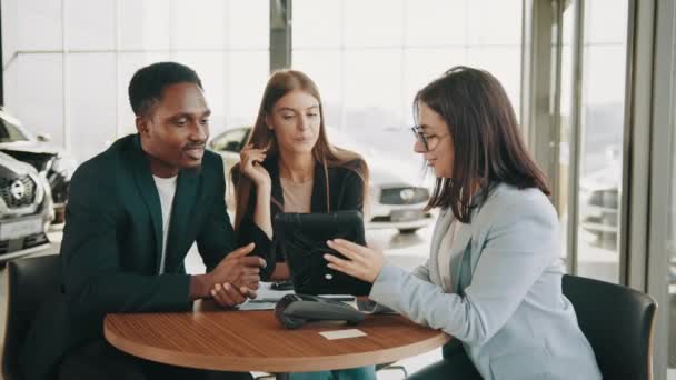 African man and caucasian woman talking with female car dealer at auto salon while making expensive purchase. Couple choosing new vehicle together. Saleswoman using tablet while working with clients — Vídeos de Stock