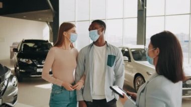 Çok ırklı maskeli çift, galeride yeni bir araba seçerken kadın satıcıyla konuşuyor. Ellerinde tabletle yetkili yönetici müşterilere danışmanlık yapıyor. Modern arabalar