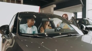 Modern lüks arabalarda oturan ve iç mekanların detaylarını inceleyen çok kültürlü güzel bir çift. Araba galerisinde aile aracı seçiyor. Satıcı kadın, sergi salonundaki çift arabalarına gösteriyor.