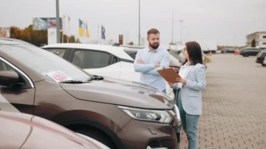 Erkek müşteri, galeride araba seçerken becerikli bir satış elemanıyla sohbet ediyor. Kadın ve erkek dışarıda lüks otomobillerin yanında dikiliyorlar. Kadın satıcı erkek müşterinin araba vurmasına yardım ediyor.