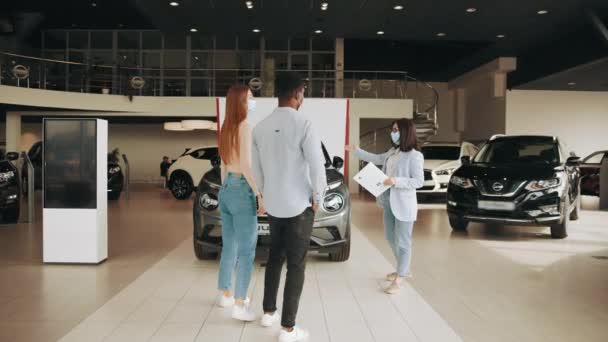 女性マネージャーの助けを借りて、現代のサロンで新しい車を選択顔マスクのアフリカ系アメリカ人男性。クライアントの高級自動車を作るために示す手のクリップボードと白人マネージャー. — ストック動画