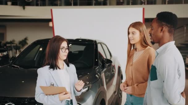 Female seller working with multiracial couple at modern auto center. Happy african man and caucasian woman consulting with manager while choosing new vehicle. Multi ethnic couple and saleswoman — Vídeos de Stock
