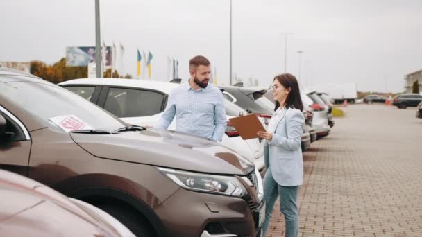 Verkoopster toont iets op klembord aan mannelijke klant van dealercentrum. Gelukkig bebaarde man koopt moderne auto met banner te koop op de voorruit. Vrouwelijke verkoper helpen mannelijke klant shoosing auto — Stockvideo