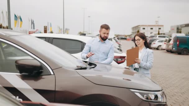 Verkäuferin zeigt männlichen Kunden des Autohauses etwas auf Klemmbrett. Glücklicher bärtiger Mann, der ein modernes Auto mit Verkaufsbanner an der Windschutzscheibe kauft. Mann spricht mit Verkäuferin über Auto im Freien — Stockvideo