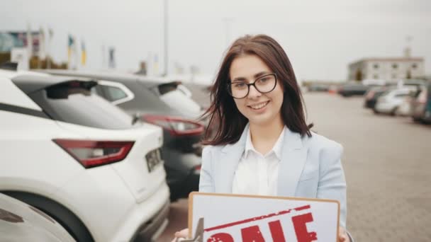 Усміхнена біла жінка з темним волоссям тримає в руках рекламний банер, стоячи на відкритому повітрі серед розкішних автомобілів. Гарна пропозиція для клієнтів. Концепція автобізнесу . — стокове відео