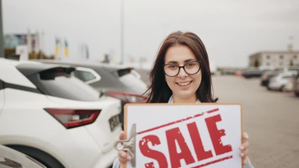 Smiling caucasian woman with dark hair holding sale banner in hands while standing outdoors among luxury cars. Good offer for clients. Auto business concept. Woman standing near row of new car — 비디오