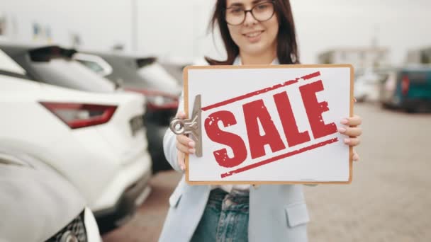 Närbild av vacker ung kvinna med försäljning banner i händerna poserar utomhus nära nya lyxbilar. Begreppet försäljning, auto business och bra erbjudande. — Stockvideo