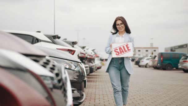 Leende kvinna med försäljning banner står bland lyxbilar. Begreppet försäljning, auto business och bra erbjudande. Kvinna stående nära raden av nya bilar med försäljning banner — Stockvideo