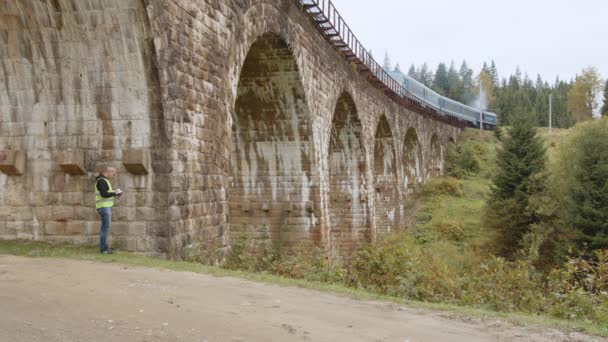Um engenheiro de cabelos grisalhos sênior está sob uma ponte ferroviária muito antiga, na qual um trem passa e faz medições. Reconstrução de antigas pontes arqueadas, preservação de antigas pontes ferroviárias — Vídeo de Stock
