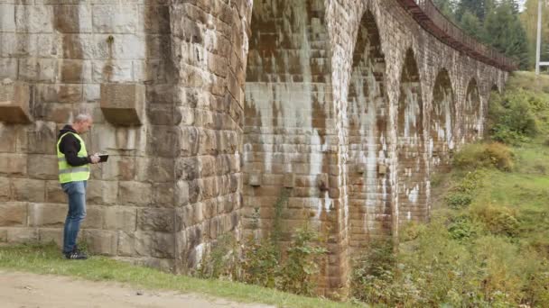 A senior gray-haired engineer stands under a very old railway bridge and takes measurements and records defects. Reconstruction of old arched bridges, preservation of ancient railway bridges — Vídeos de Stock