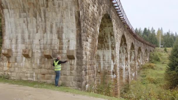 检查桥梁。一位头发灰白的高级工程师站在一座非常古老的铁路桥下，测量并记录缺陷。旧桥的重建、古桥的保护 — 图库视频影像