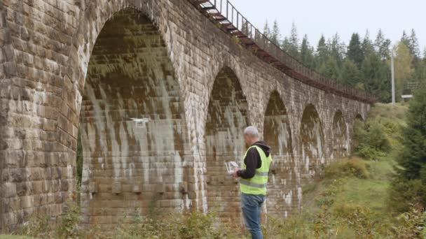 Um drone debaixo de uma ponte a fazer uma inspecção. Operador de drones ao lado do viaduto. Quadcopter para observação de ponte. Tecnologias de manutenção de estradas e pontes. Engenheiro com drone. Controlo aéreo — Vídeo de Stock