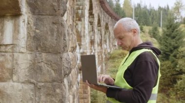 Eski binaları ve köprüleri restore etmek için kıdemli erkek mühendis. Mühendis veriyi dizüstü bilgisayara yazar. Köprülerin yeniden inşası, antik demiryolu köprülerinin korunması. Mühendis müfettişi iş başında.