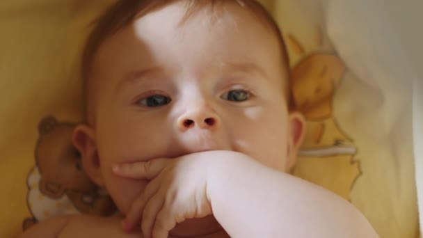 Porträt eines verspielten und energiegeladenen Kindes aus nächster Nähe, das einen Finger lutscht. Ein nettes kleines kaukasisches Neugeborenes lächelt lustig. Baby lächelt lustig und liegt hinten in der Kinderkrippe — Stockvideo