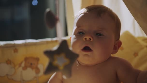 Um bebê rindo brincando com brinquedos de pendurar celular e olhando para a câmera. Cuidados com o bebé, primeiras habilidades, aprender a sentar-se. Brinquedos para berço infantil. Criança sorridente brincando com urso de brinquedo no quarto ensolarado. — Vídeo de Stock