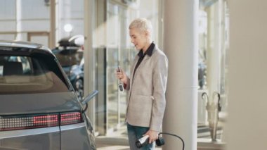 Eko elektrikli araba konsepti. Kadın elektrikli aracına doğru yürüyor, park yerinde şarj etmek için fişe takıyor. İş kadını, kablolu TV 'yi yeni elektrikli arabasına bağlıyor. İş merkezinde hızlı şarj oluyor.