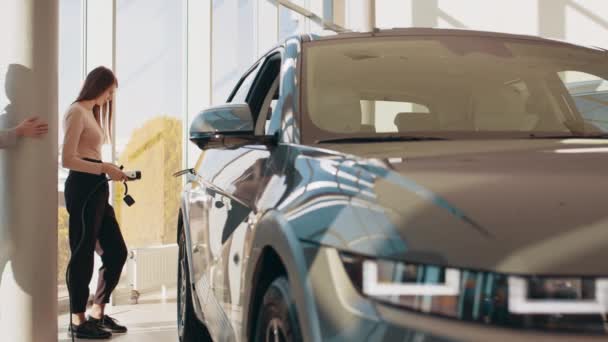 Duas jovens mulheres bonitas felizes estão cobrando um carro elétrico pela primeira vez em uma estação de carregamento em um salão ou escritório moderno. O conceito de transporte amigo do ambiente. Carro elétrico Eco — Vídeo de Stock