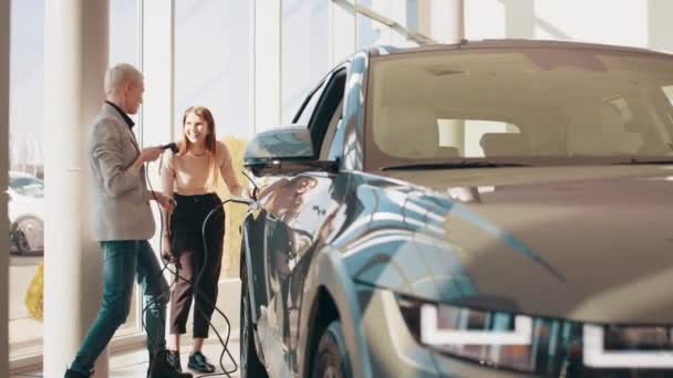 Dos mujeres hermosas jóvenes felices están cargando un coche eléctrico por primera vez en una estación de carga en un salón moderno u oficina. El concepto de transporte respetuoso con el medio ambiente. Eco coche eléctrico — Vídeos de Stock