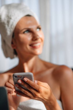 Kafkasyalı bir kadının banyo havlusuna sarılıp mobil ekrana bakması ve mutluluğu ifade etmesi. İyi haber, duygular ve teknoloji kavramı.
