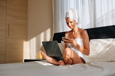 Banyo havlusuna sarılı güzel bir kadın dizüstü bilgisayarla yatağa uzanmış ve akıllı telefonu elinde tutuyor. Modern teknolojili yaşam tarzları. Bir kadın banyodan ya da duştan sonra otelde tatilde çalışır..