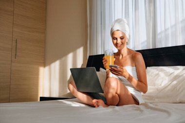 Banyo havlusuna sarılmış bir kadın kucağında dizüstü bilgisayarla yatağa uzanmış ve elinde bir akıllı telefon tutuyordu. Modern teknolojilerle yaşam tarzı. Bilgisayarlı genç bir kız telefonda konuşuyor ve gülümsüyor.