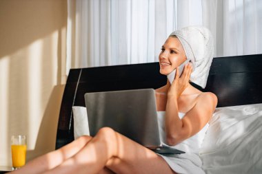 Banyo havlusuna sarılı güzel bir kadın dizüstü bilgisayarla yatağa uzanmış ve akıllı telefonu elinde tutuyor. Modern teknolojili yaşam tarzları. Bir kadın banyodan ya da duştan sonra otelde tatilde çalışır..