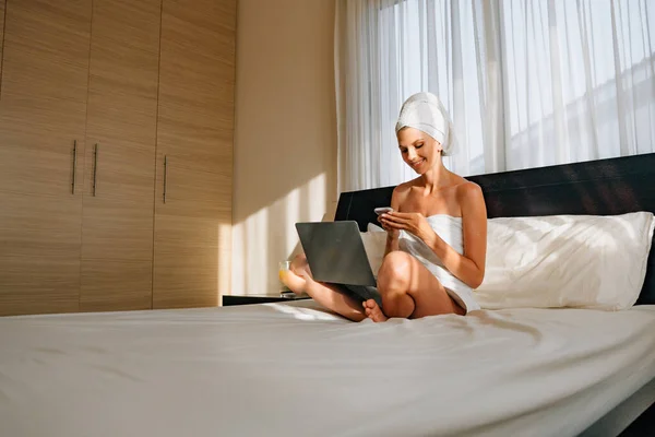 Nainen Käärittynä Kylpypyyhkeeseen Makaa Sängyssä Kannettava Tietokone Sylissään Pitää Älypuhelinta — kuvapankkivalokuva