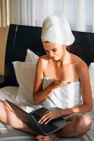 Nainen Käärittynä Kylpypyyhkeeseen Makaa Sängyssä Kannettava Tietokone Sylissään Pitää Älypuhelinta — kuvapankkivalokuva