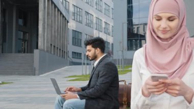 Tahta bankta oturan ve modern cep telefonu tutan tesettürlü hoş bir kadın. Kablosuz dizüstü bilgisayarda çalışan Arap bir adamın bulanık geçmişi. İş adamları ve aletler kavramı