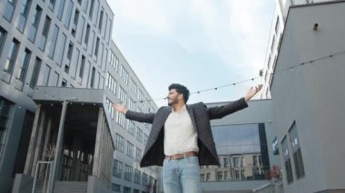 Modern ofis binasının yanında uzanan ve mutluluğu hisseden yakışıklı Hindu işadamının düşük açılı görüntüsü. İnsani duygular ve başarı kariyeri kavramı. Mutlu Hindu işadamı.